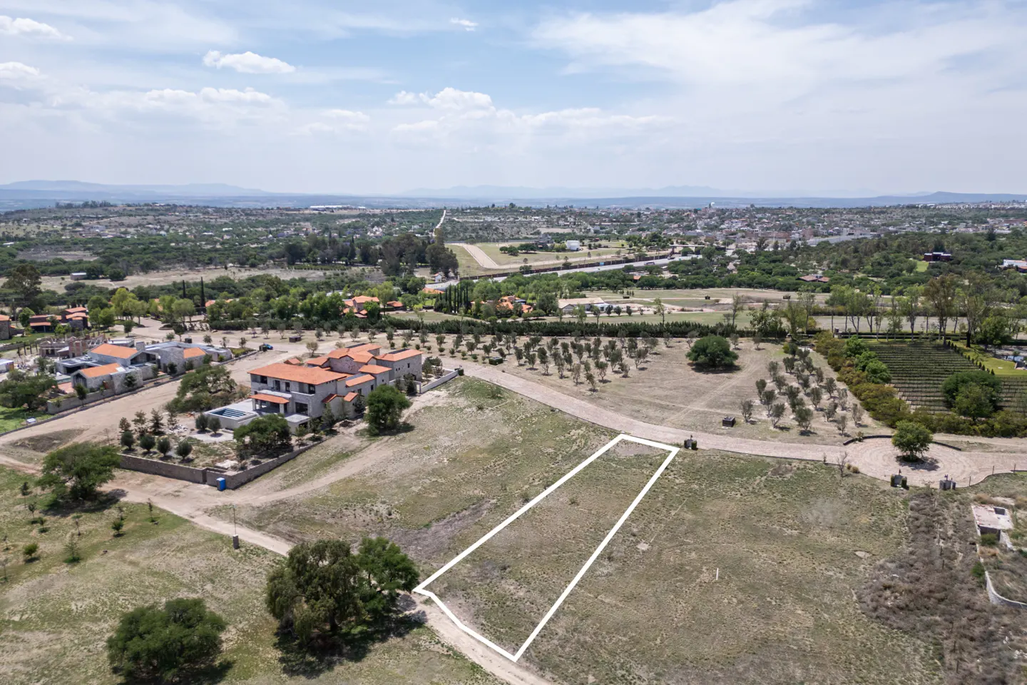 Aerial view of a vacant lot outlined in white, with luxury homes and a city skyline in the background.