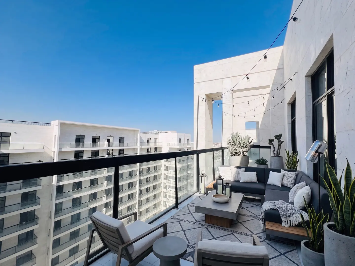 Luxury condo balcony with outdoor seating, plants, and string lights. City view with blue sky.