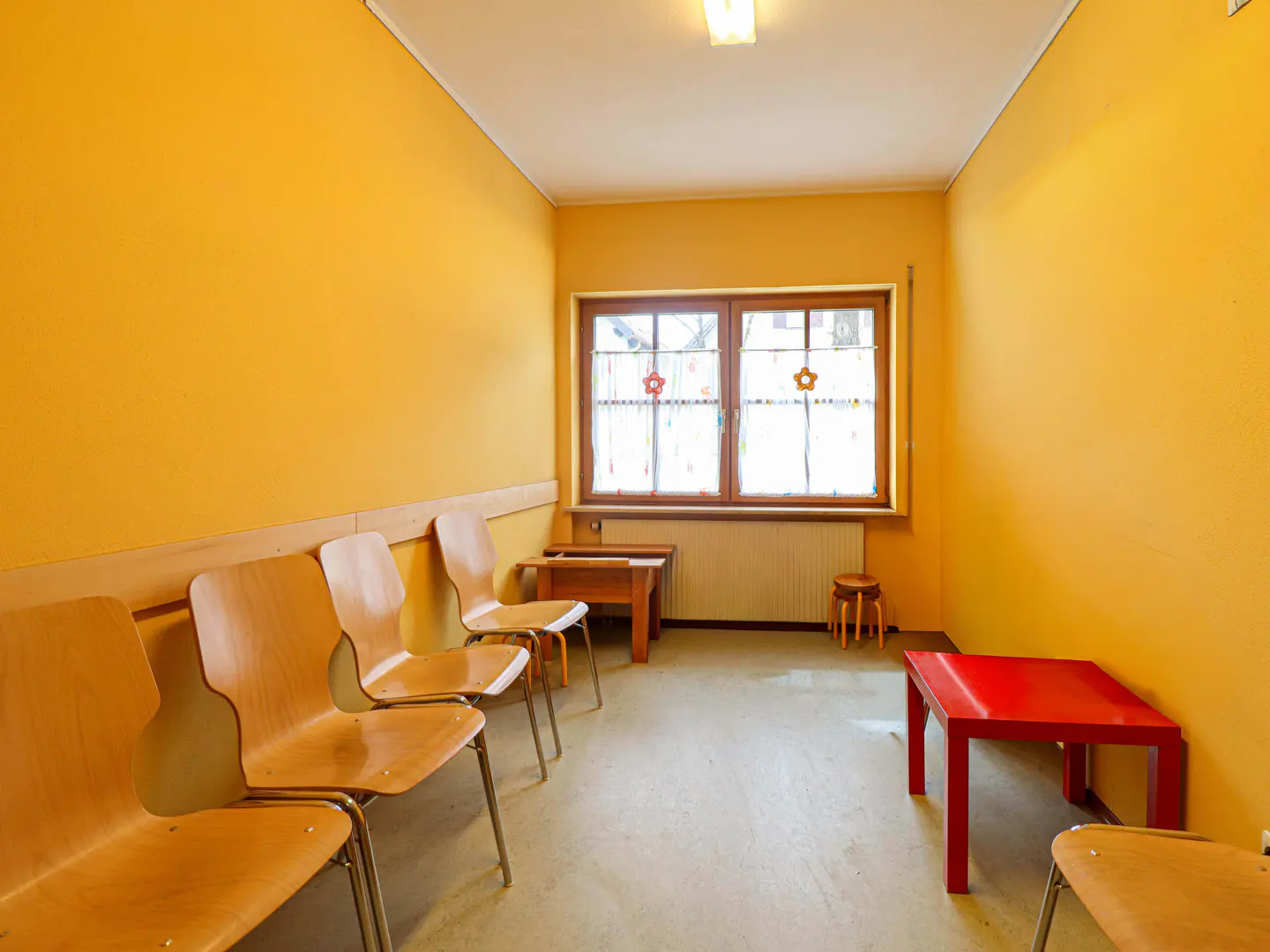 Waiting room with yellow walls, wooden chairs, and a red table. A window with flower curtains is visible.