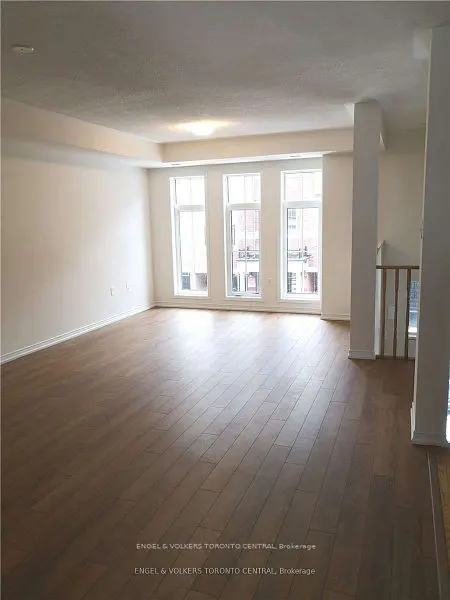 Bright, empty room with hardwood floors, white walls, and three large windows. A staircase is visible on the right.