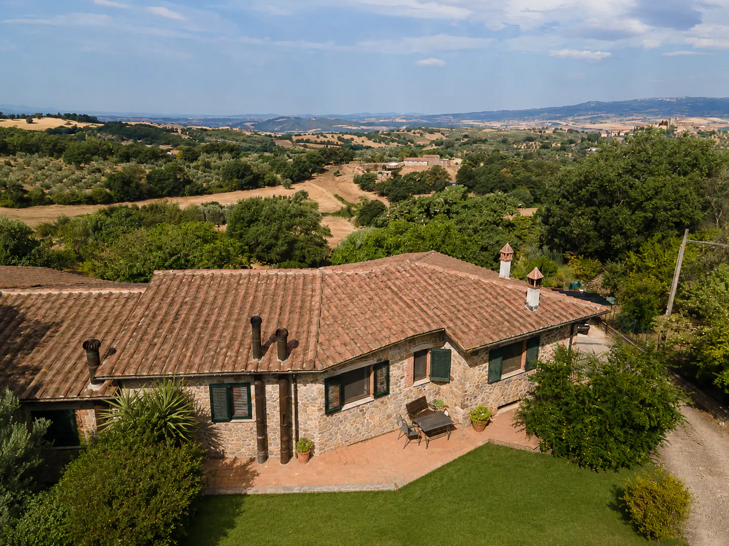 Aerial view of a stone house with a red tile roof, green shutters, and a lawn, set in a lush, hilly landscape.