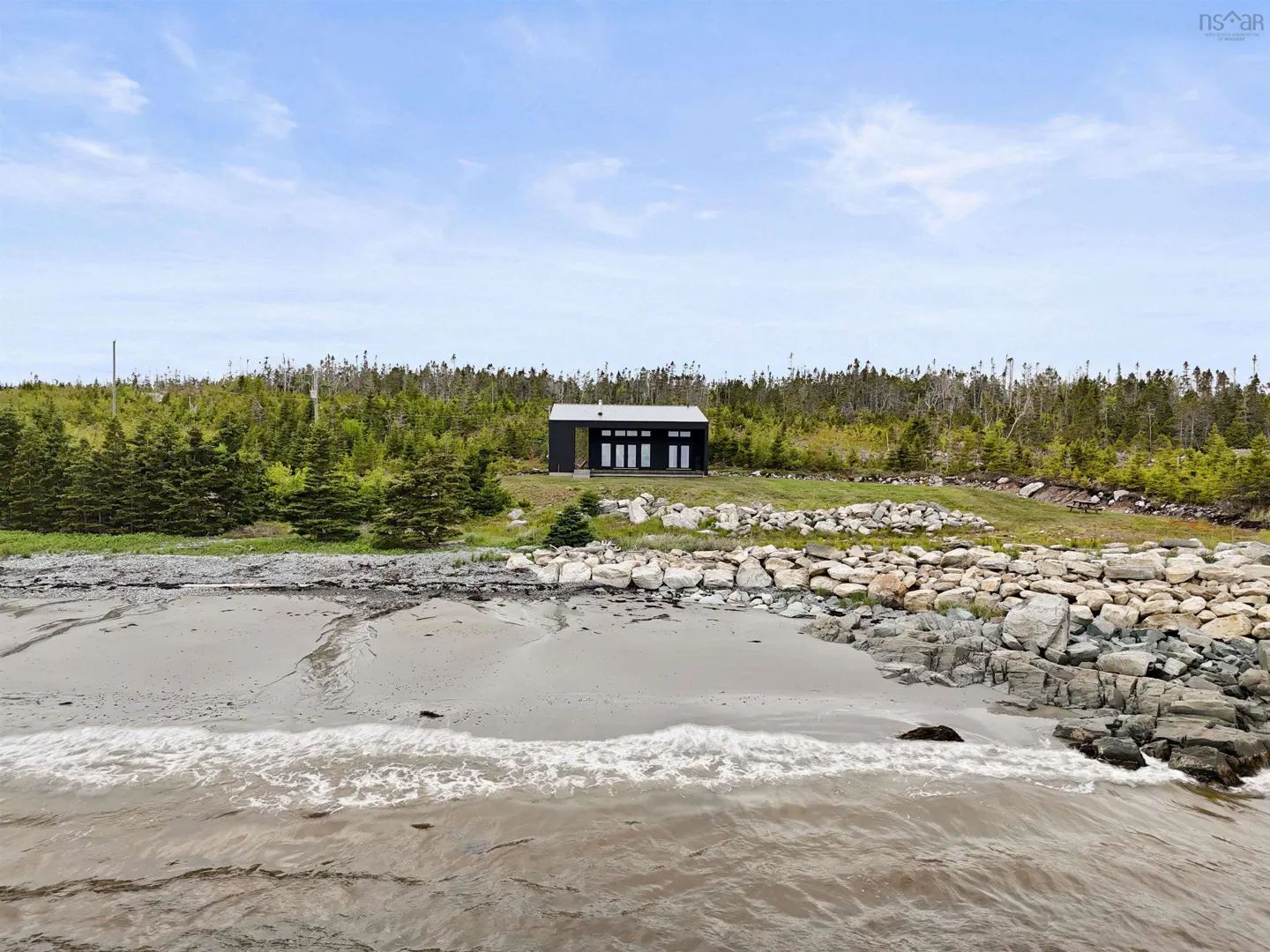 Modern black house on a grassy lot overlooking a rocky beach and the ocean under a bright blue sky.