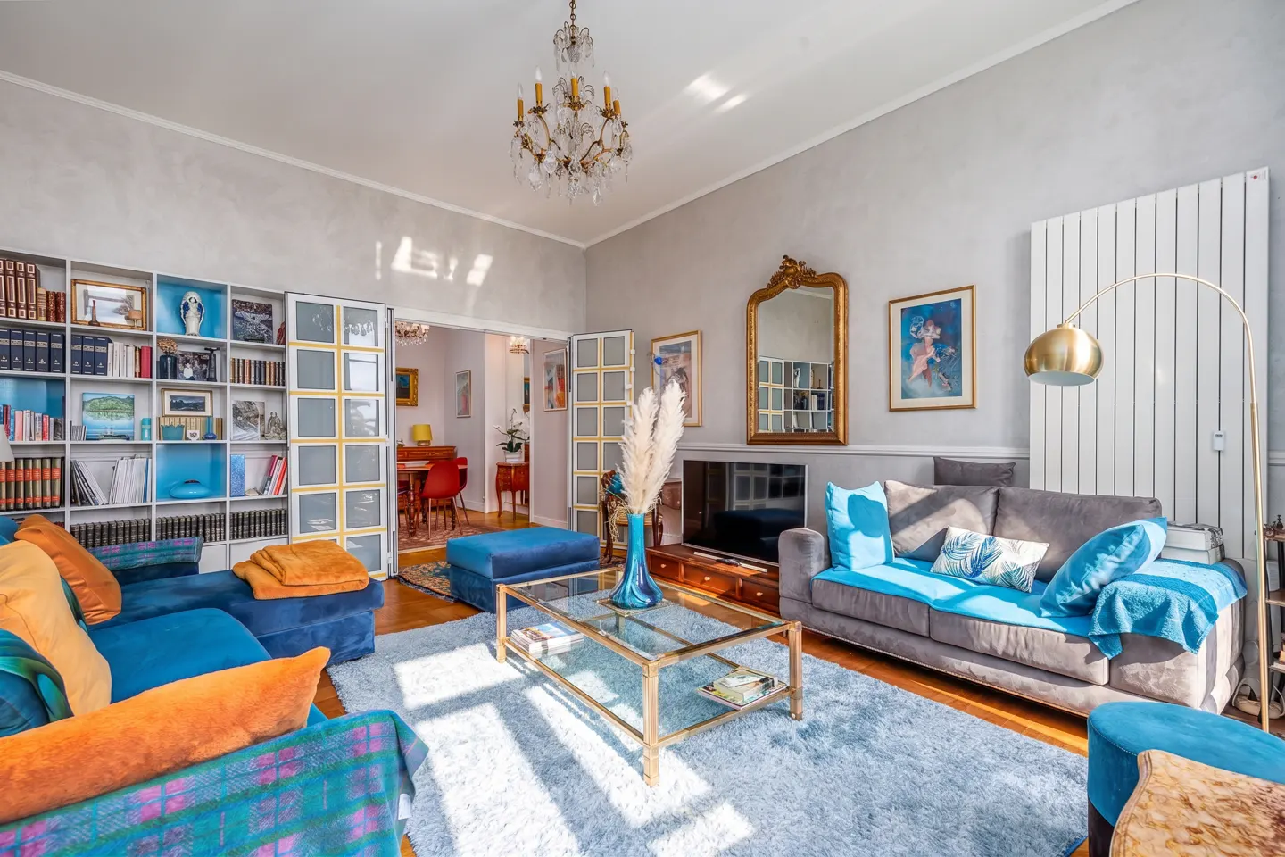 Bright living room with blue and orange sofas, a glass coffee table, a crystal chandelier, and a large wall bookshelf.