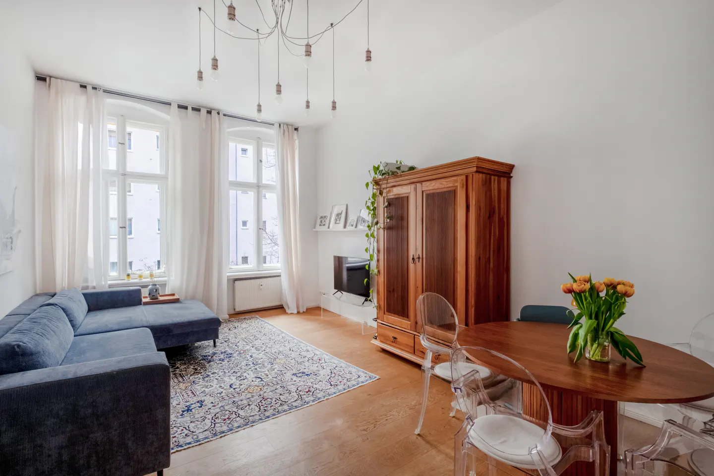 Bright living room with a blue sectional sofa, patterned rug, wood dining table with clear chairs, and a large wooden armoire. White walls and curtains.