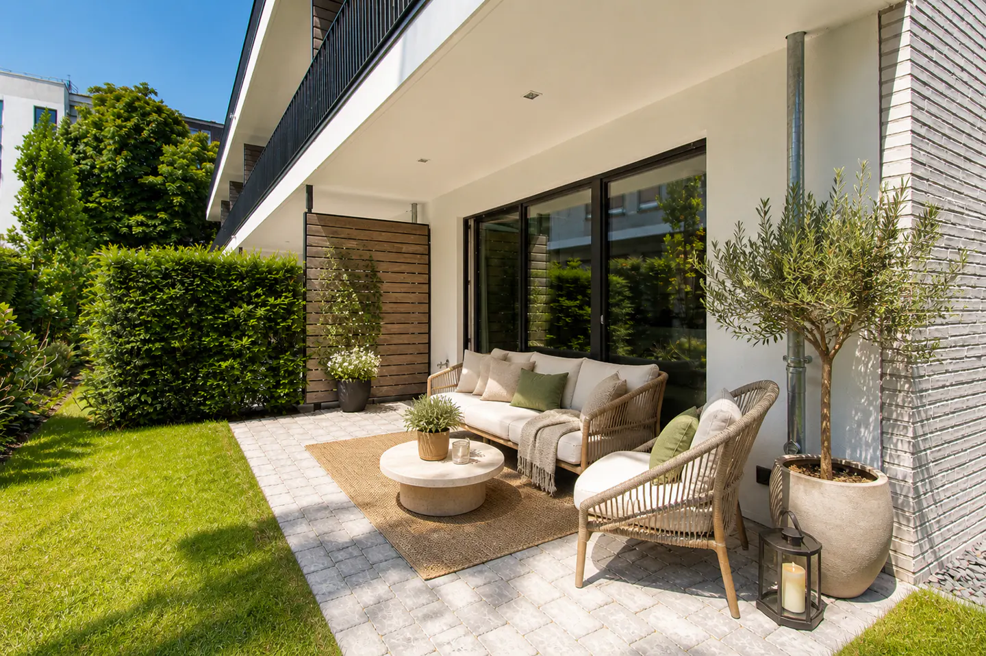 Outdoor patio with wicker furniture, including a sofa, chair, and round table on a jute rug. A potted olive tree and green lawn add to the serene setting.
