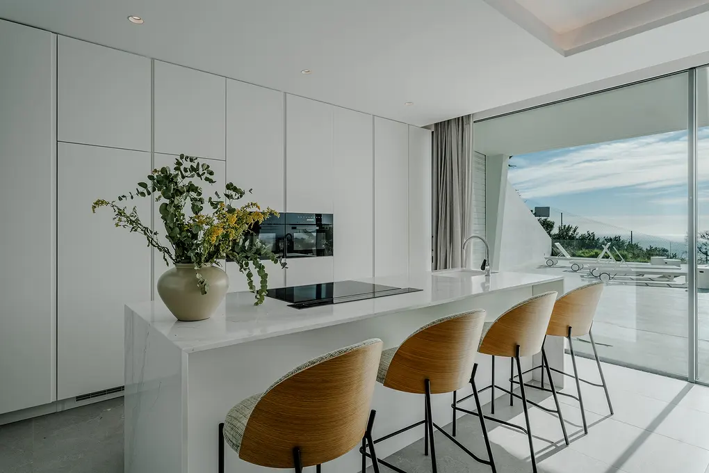 Bright, modern kitchen with white cabinets, island, and four bar stools. A vase of greenery sits on the island. Large windows offer a view of the outdoors.