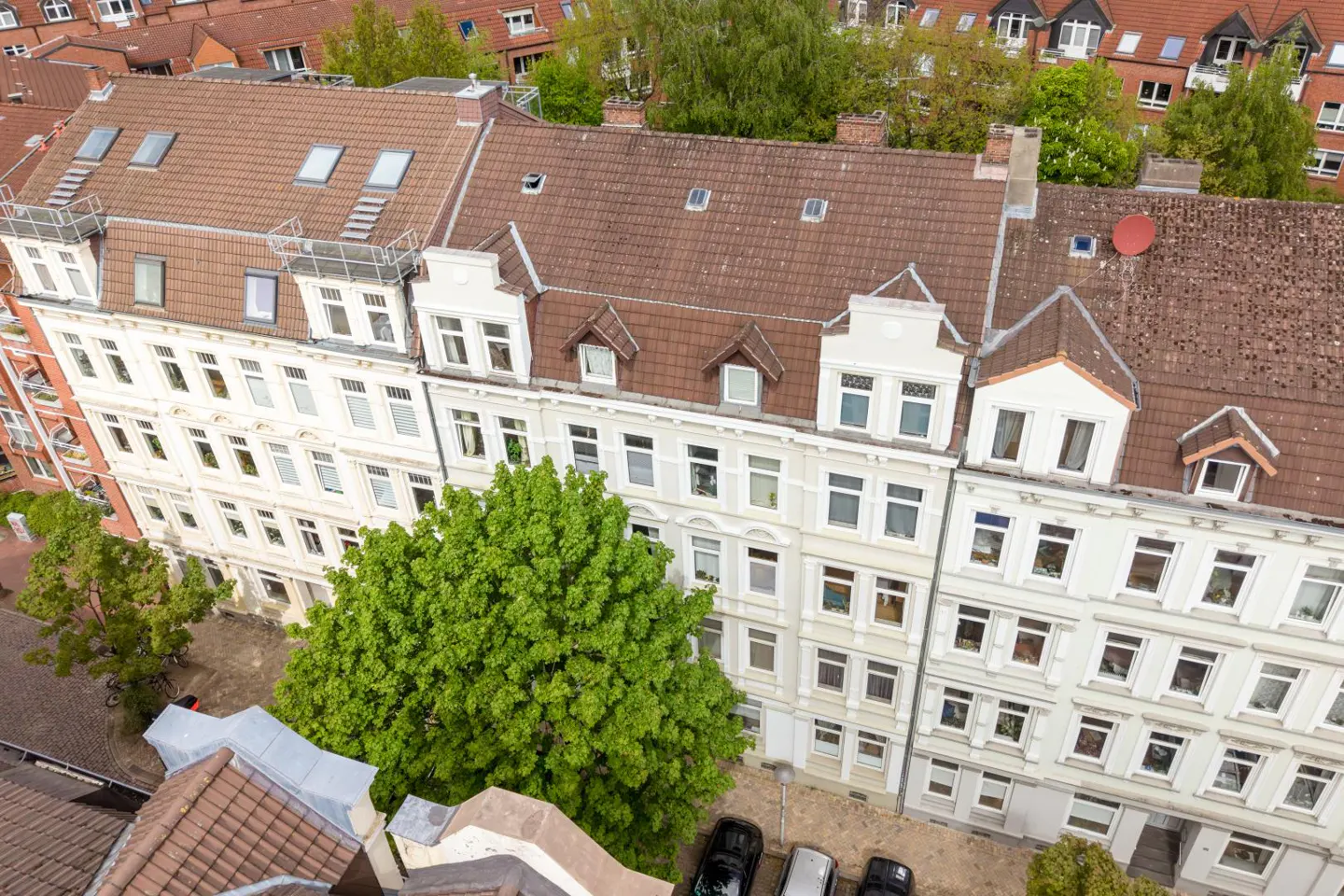 Aerial view of multi-story white apartment buildings with brown roofs, green trees, and parked cars below.