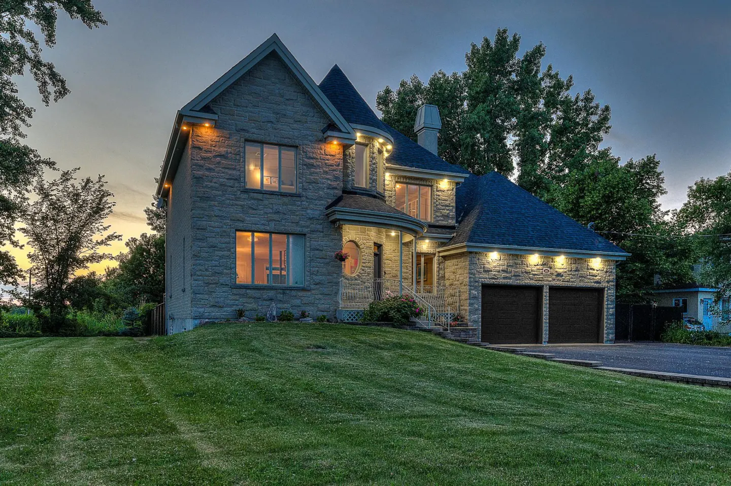 Two-story stone house with a dark roof, a two-car garage, and a green lawn at dusk.