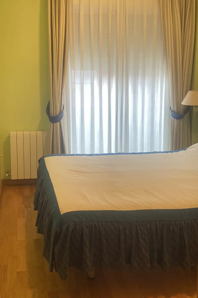 Bedroom with light green walls, wood floors, a bed with a blue ruffled skirt, and sheer curtains over a window.