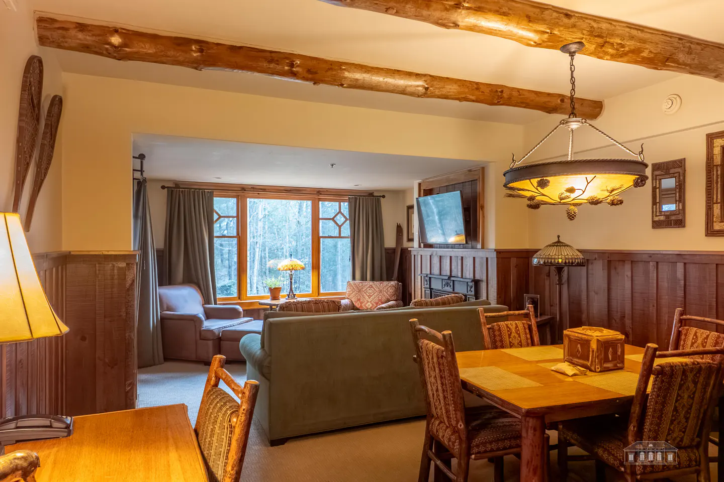 Living room with wood beams, dining table, green sofa, and a window view of trees. Snowshoes hang on the wall.
