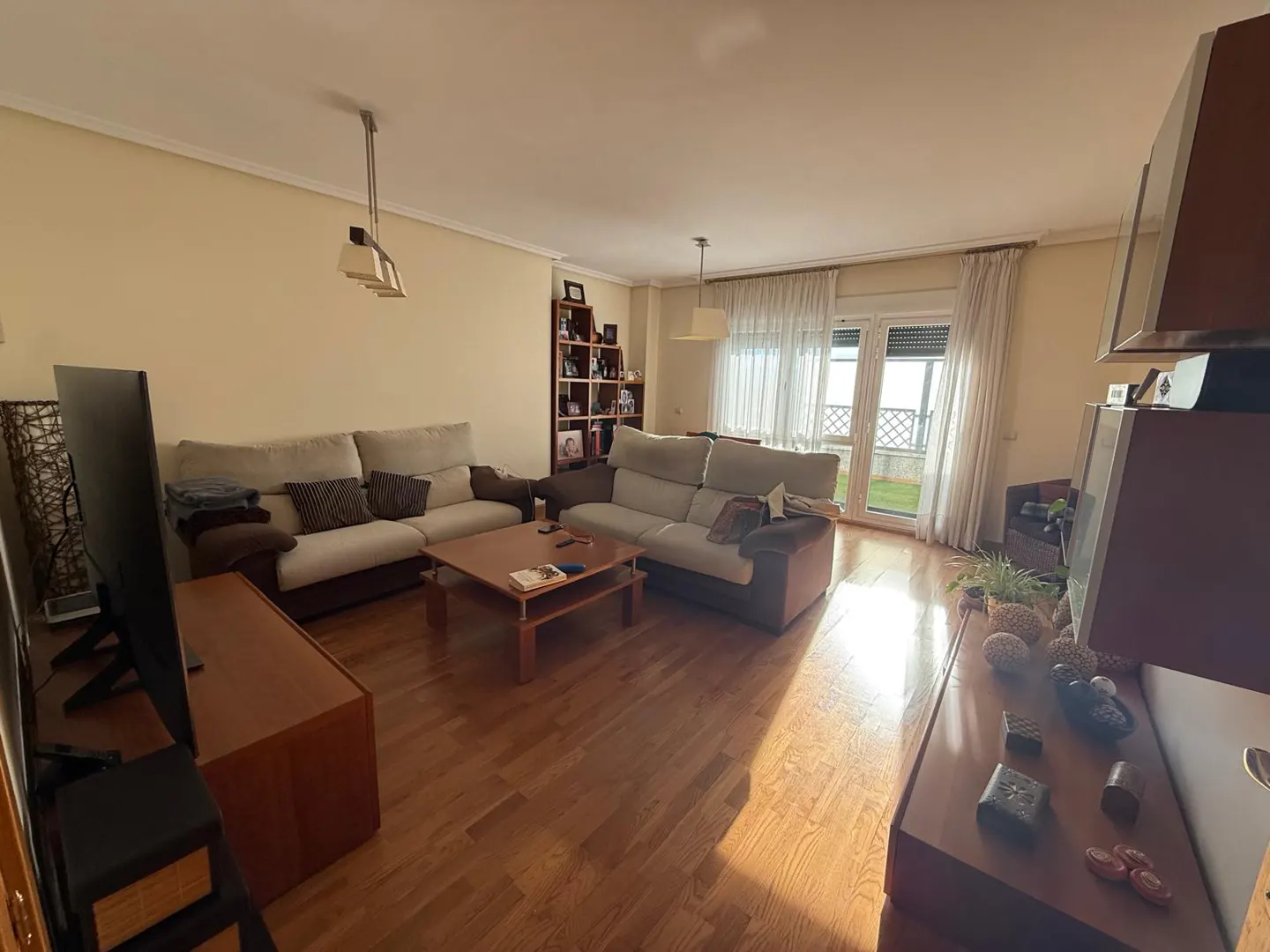 A bright living room with two sofas, a wooden coffee table, and a TV stand on a hardwood floor. A bookcase and a door to a balcony are visible.