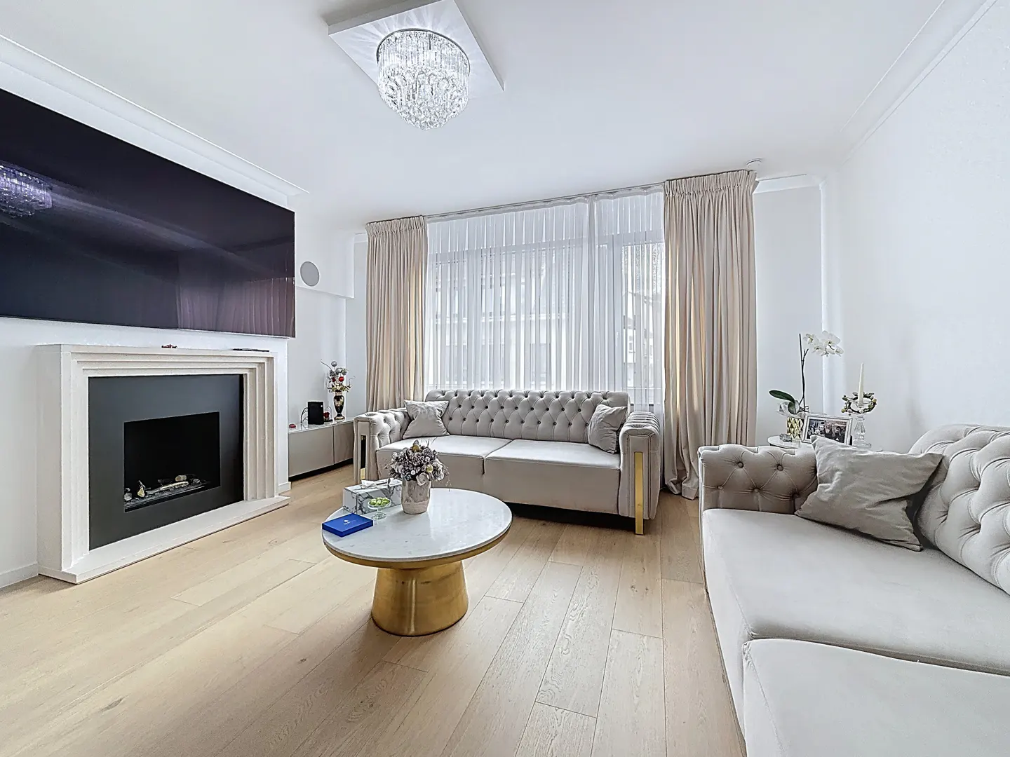 Bright living room with light wood floors, white walls, and beige sofas. A fireplace with a large TV above it is on the left. A crystal chandelier hangs from the ceiling.