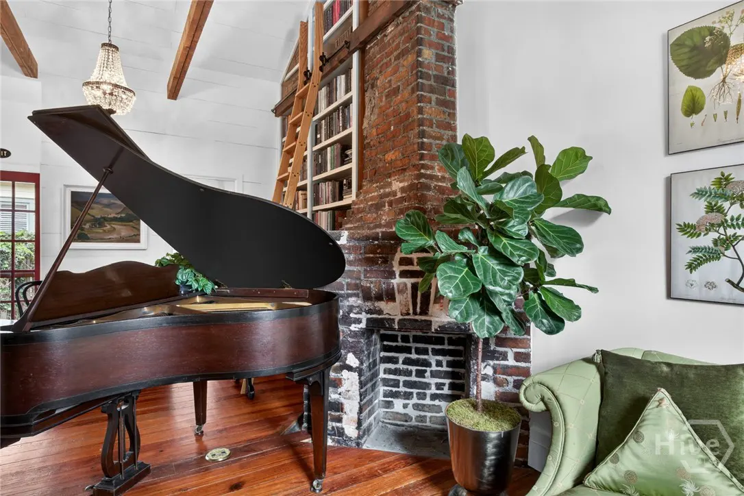 Living room with a grand piano, brick fireplace, and a bookshelf with a ladder. A chandelier hangs from the ceiling.