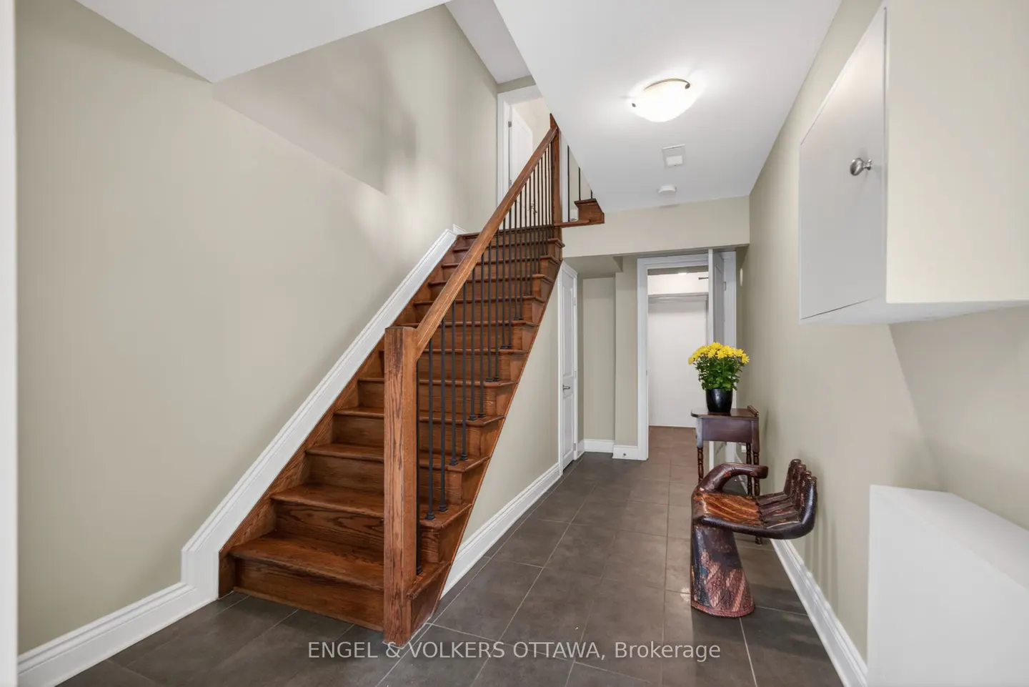 Interior view of a hallway with a wooden staircase, brown tile floor, beige walls, and a decorative wooden chair.