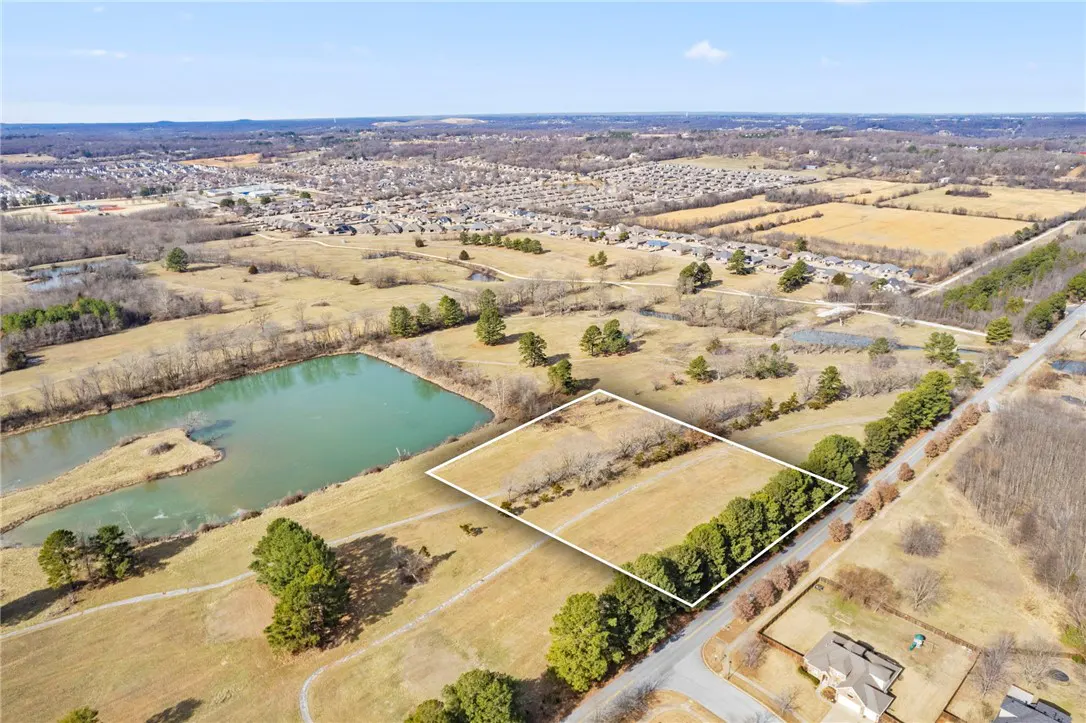 Aerial view of a vacant lot outlined in white, near a pond and residential area under a blue sky.