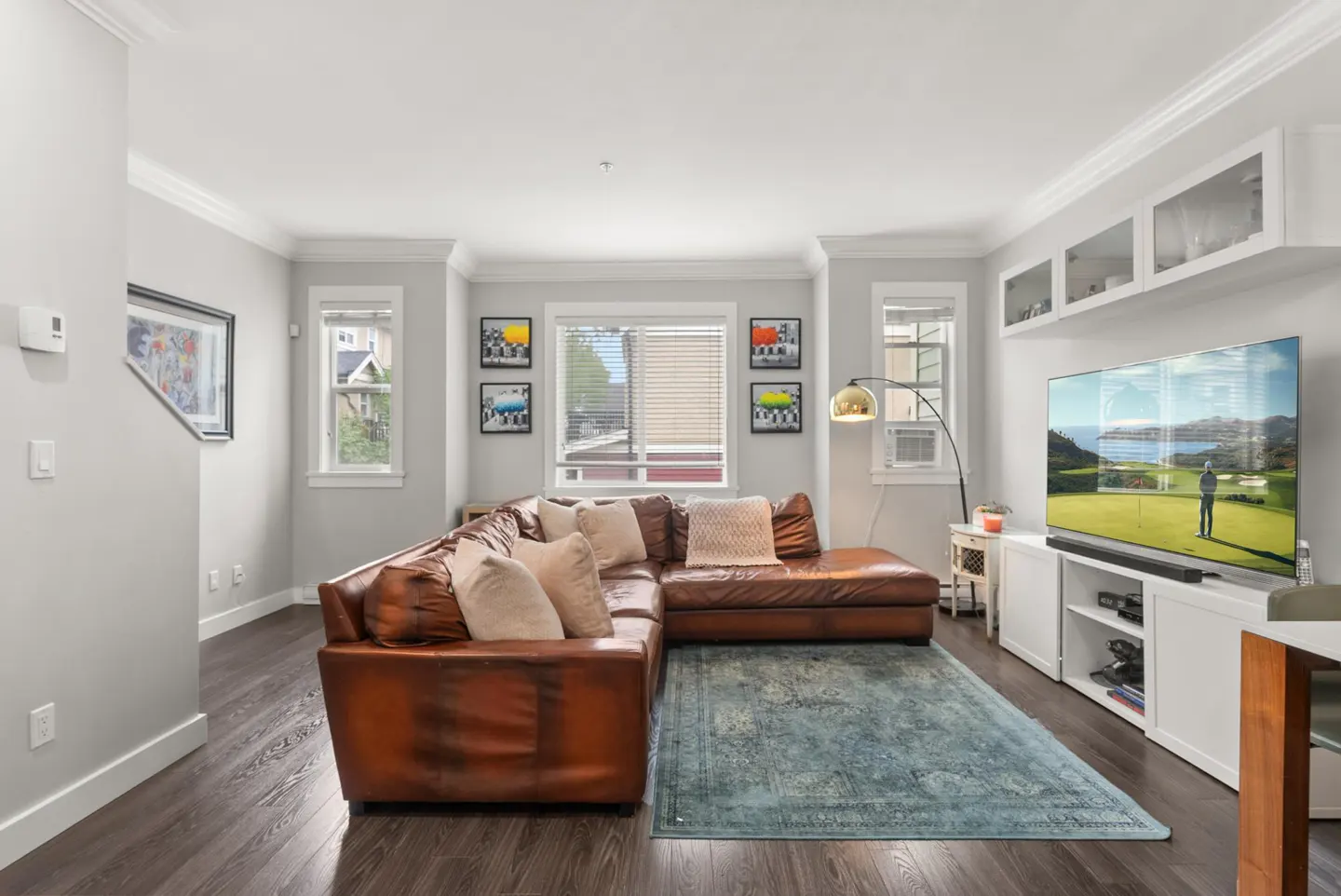 Living room with brown leather sectional, blue rug, and TV showing a golf course. Gray walls and dark wood floors.