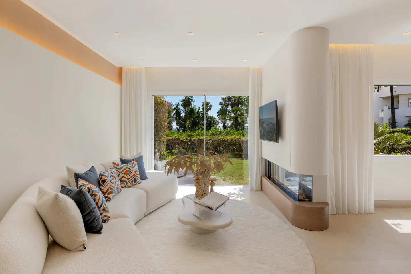Bright living room with a white curved sofa, patterned pillows, and a round rug. A modern fireplace and a large window with a garden view.
