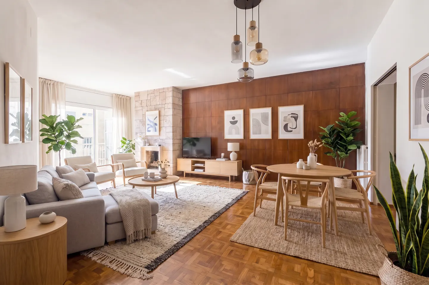 Bright living room with a gray sofa, wood floors, a stone fireplace, and a round wood dining table. Three abstract art prints hang on the wood paneled wall.