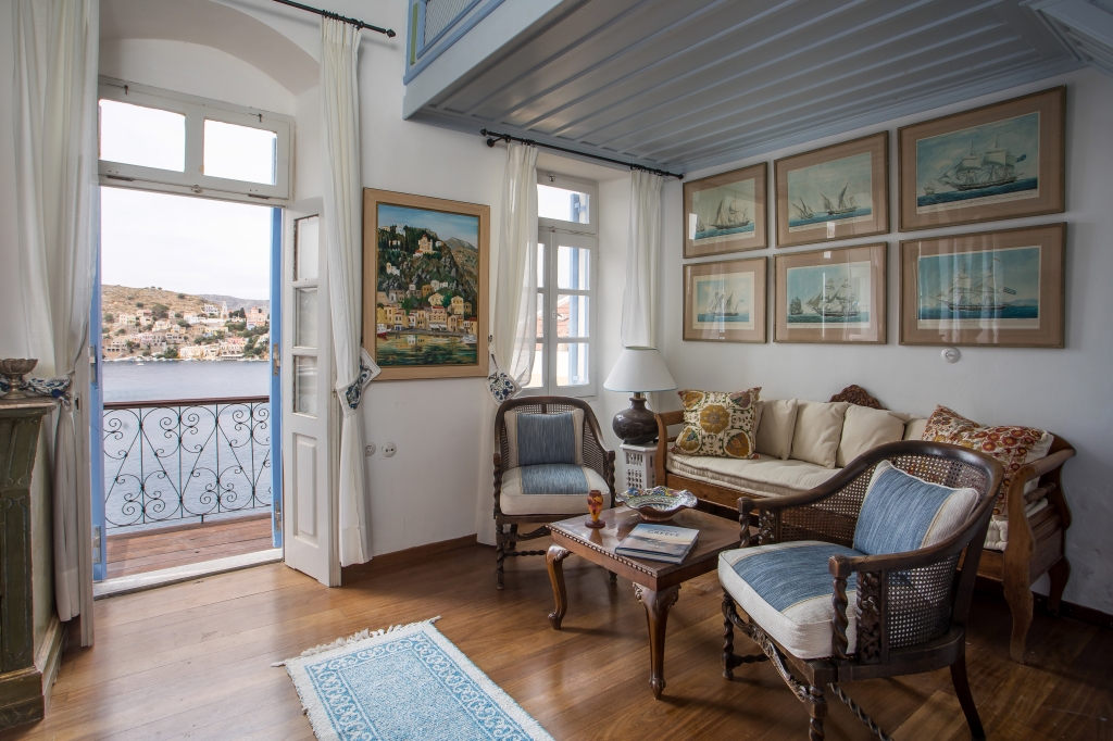 Living room with wooden floors, wicker chairs, sofa, and balcony door overlooking a coastal town and harbor.