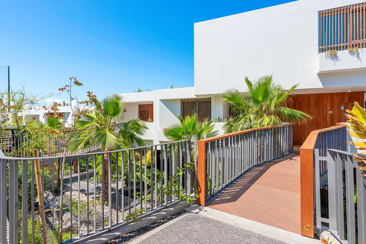 Modern white building with a wooden walkway and gray railings, surrounded by palm trees and lush greenery under a clear blue sky.