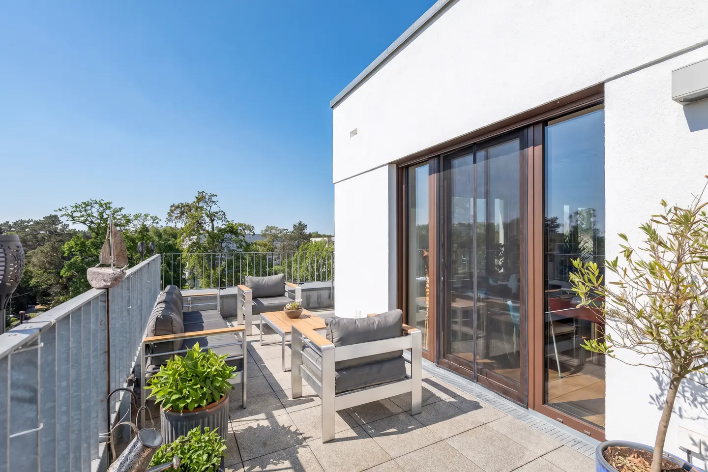 Outdoor patio with metal railing, chairs, and table. Sliding glass doors lead into a white building with trees in the background.