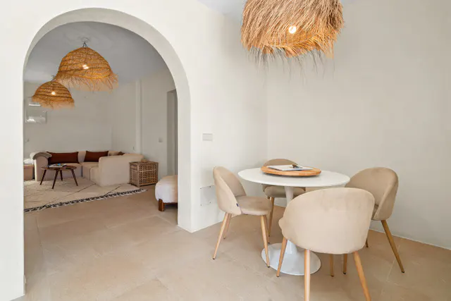 Bright, neutral living space with a round white table, four beige chairs, and straw pendant lights. A living room is visible through an arched doorway.