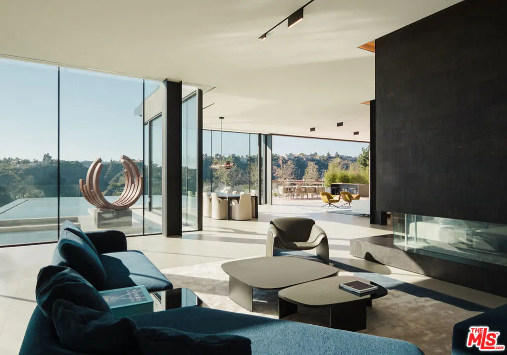 Bright, modern living room with blue sofa, dark coffee tables, and a black fireplace. Large windows overlook a pool and green landscape.