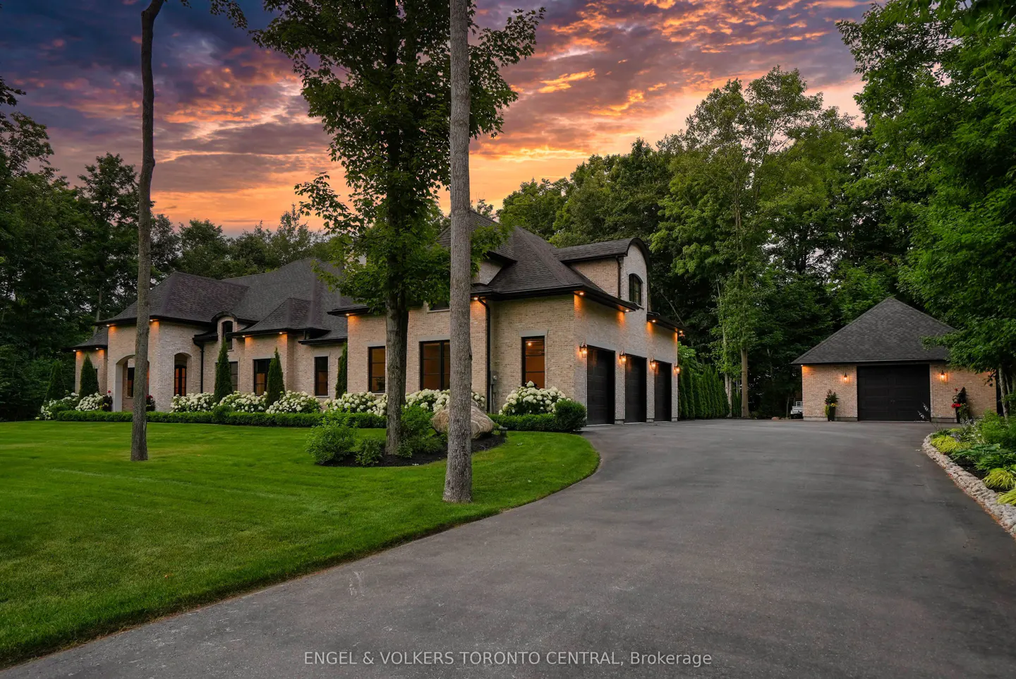 A large, tan brick house with a dark roof, three-car garage, and a long driveway at sunset.
