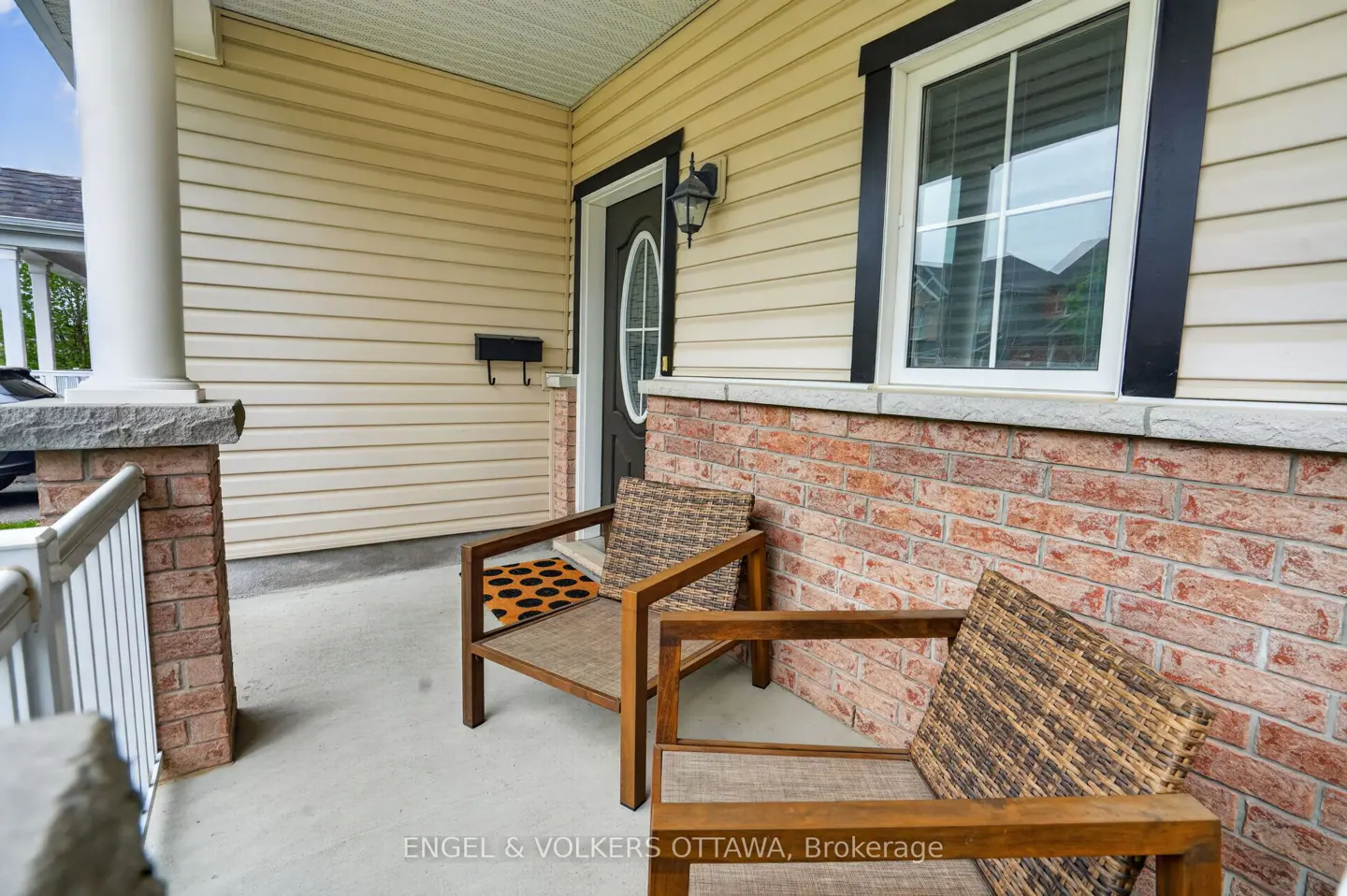 Covered porch with two wooden chairs with woven seats, brick and beige siding, black door, and window trim.
