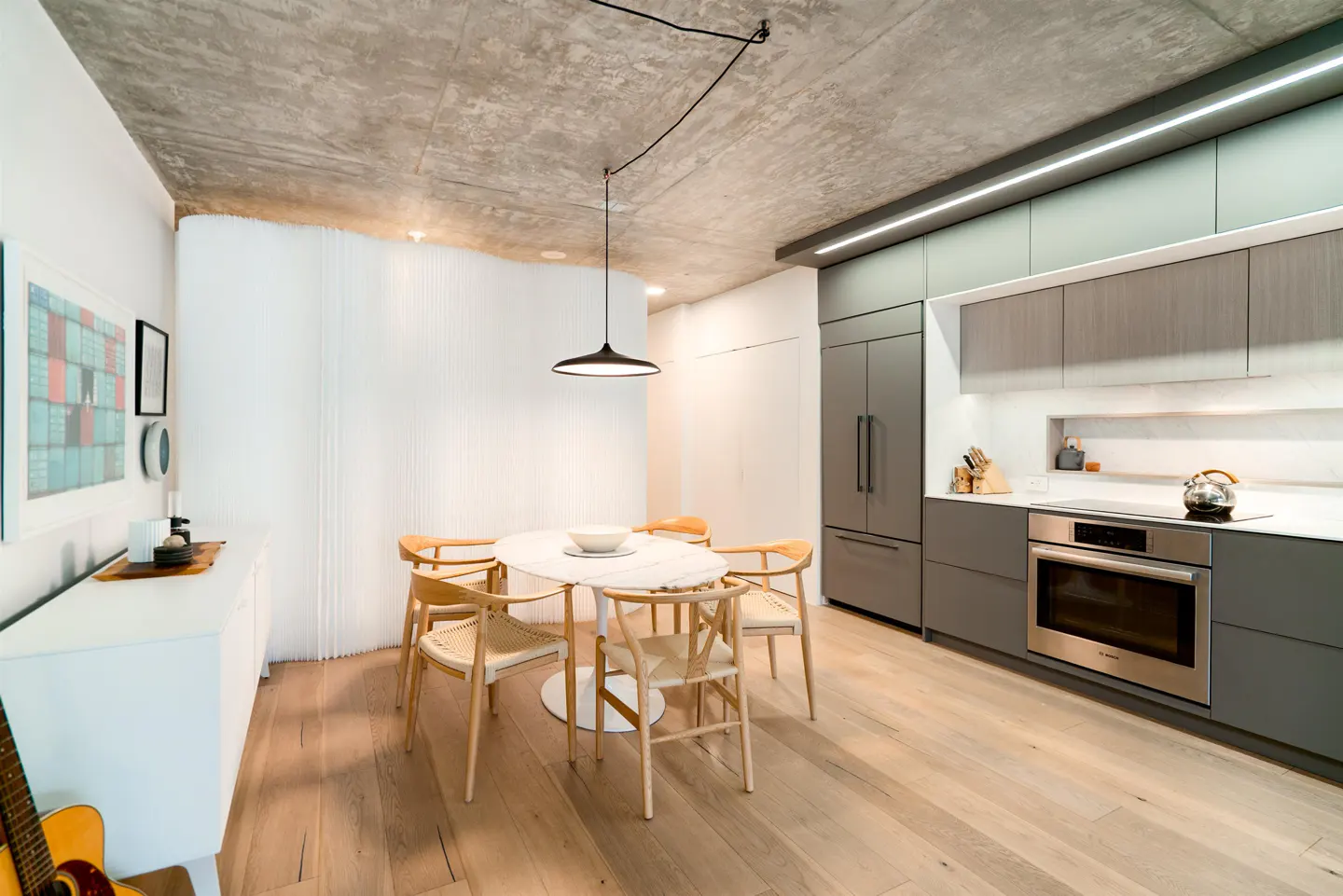 A modern kitchen with a concrete ceiling, gray cabinets, and a light wood floor. A white table with wooden chairs sits under a black pendant light.