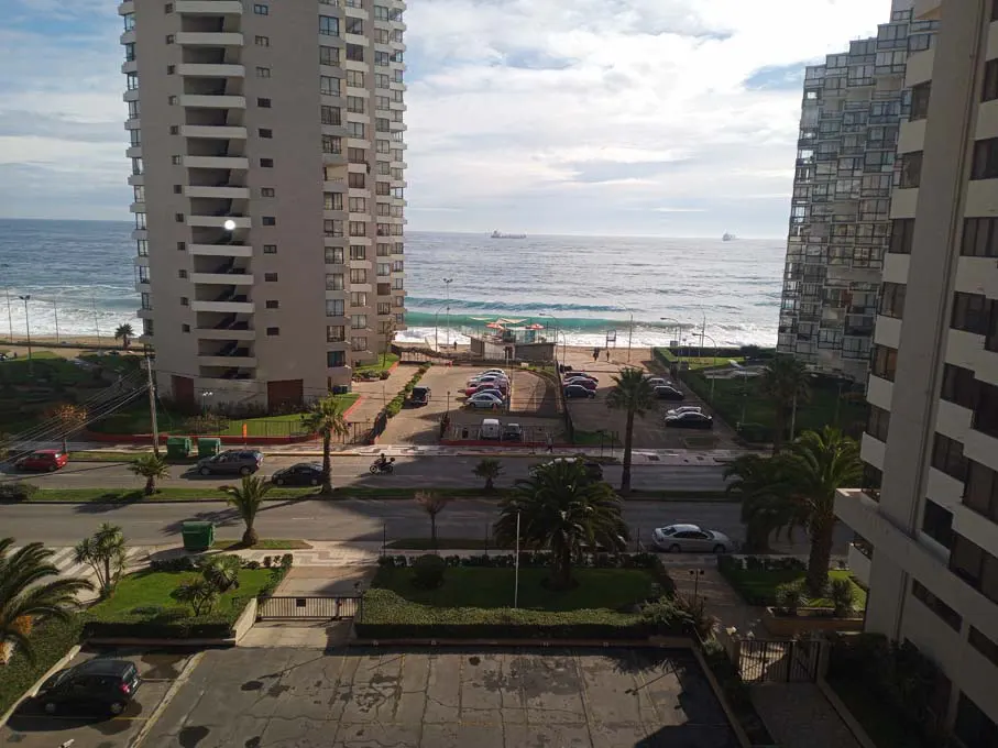 View from a high-rise apartment overlooking a beach with ocean views. Tall buildings frame the scene. Cars are parked near the beach.