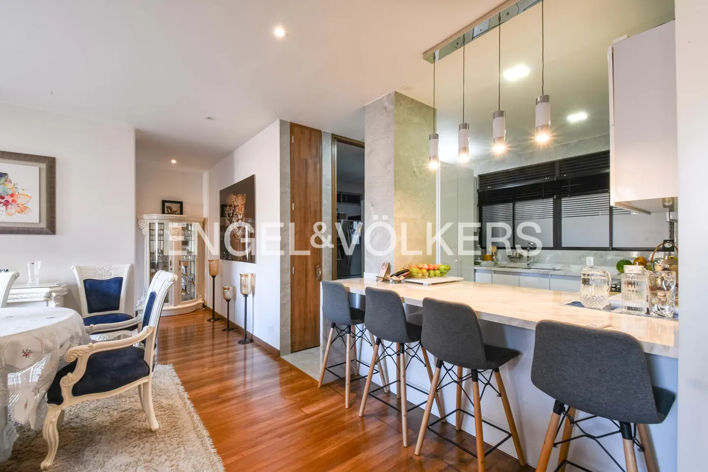 A bright, modern apartment interior with a kitchen island, gray stools, and a dining area with blue and white chairs. Hardwood floors and white walls.
