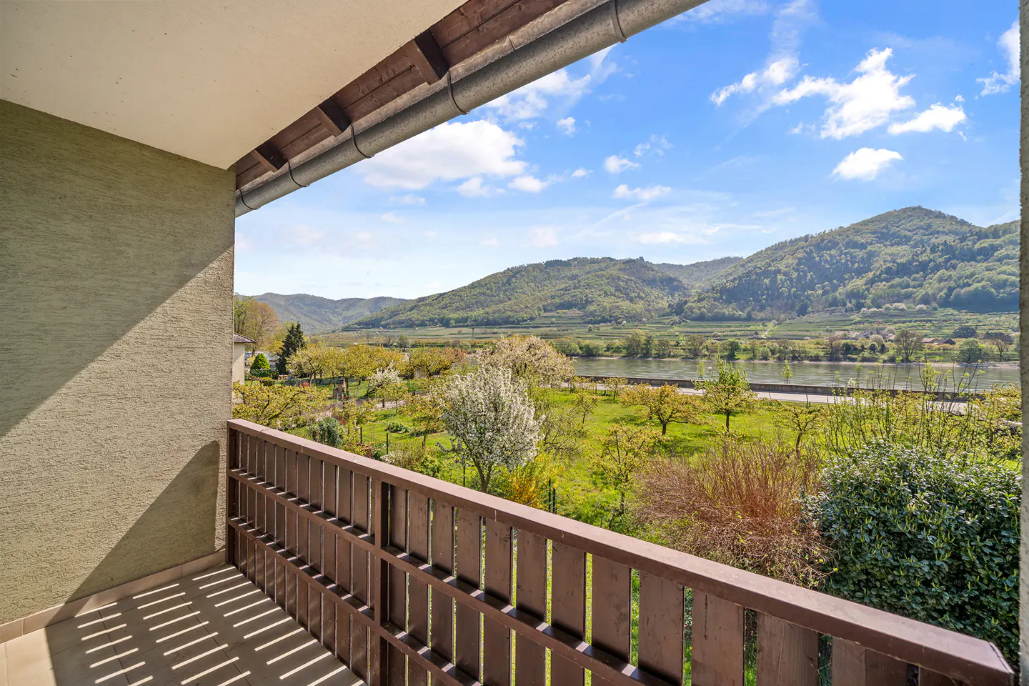 Balcony view of green hills and a river under a blue sky. Brown wooden railing in the foreground.