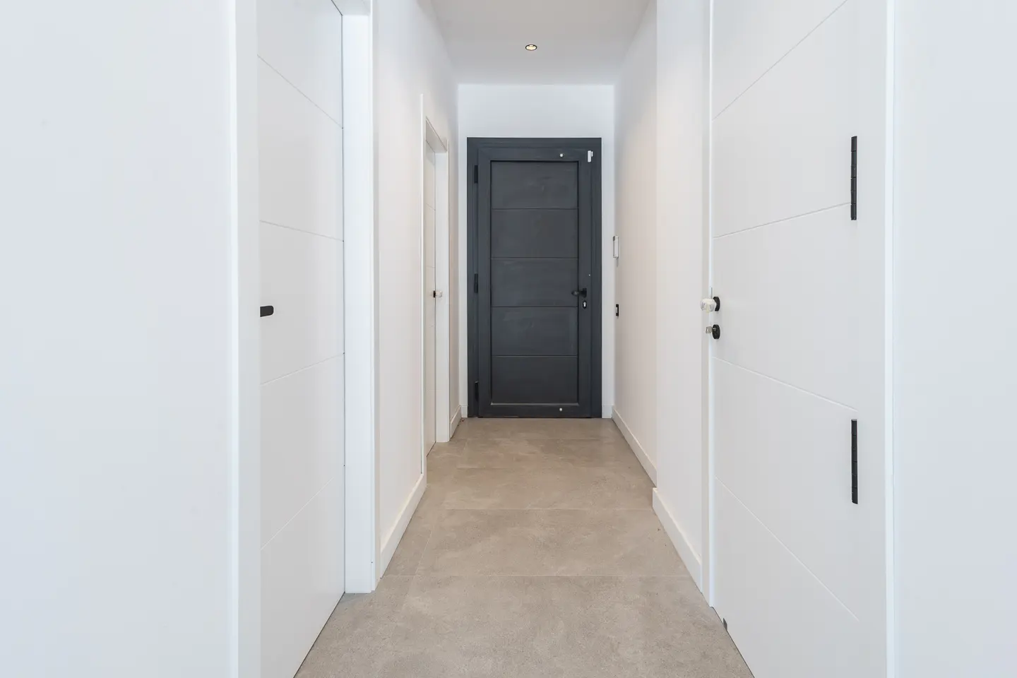 A bright hallway with white walls and doors, and a gray floor. A dark gray door is at the end of the hall.