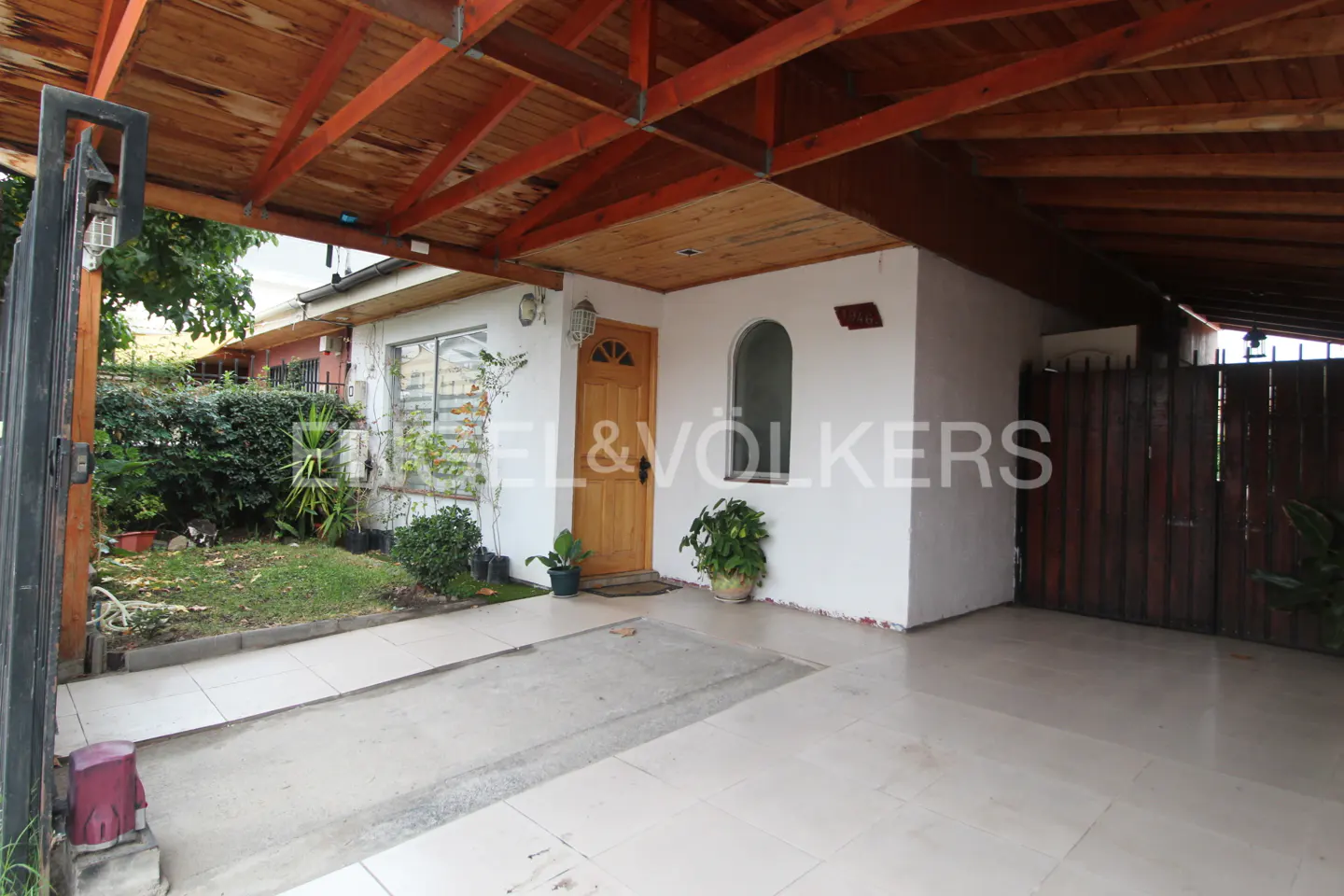 Exterior view of a white house with a wooden door and carport, a small garden, and a gated driveway.