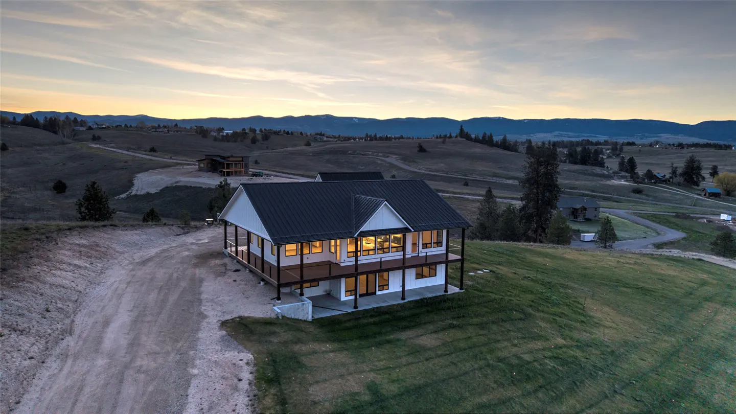Two-story white house with a black roof and a wraparound porch on a grassy hill at dusk. Mountains in the background.