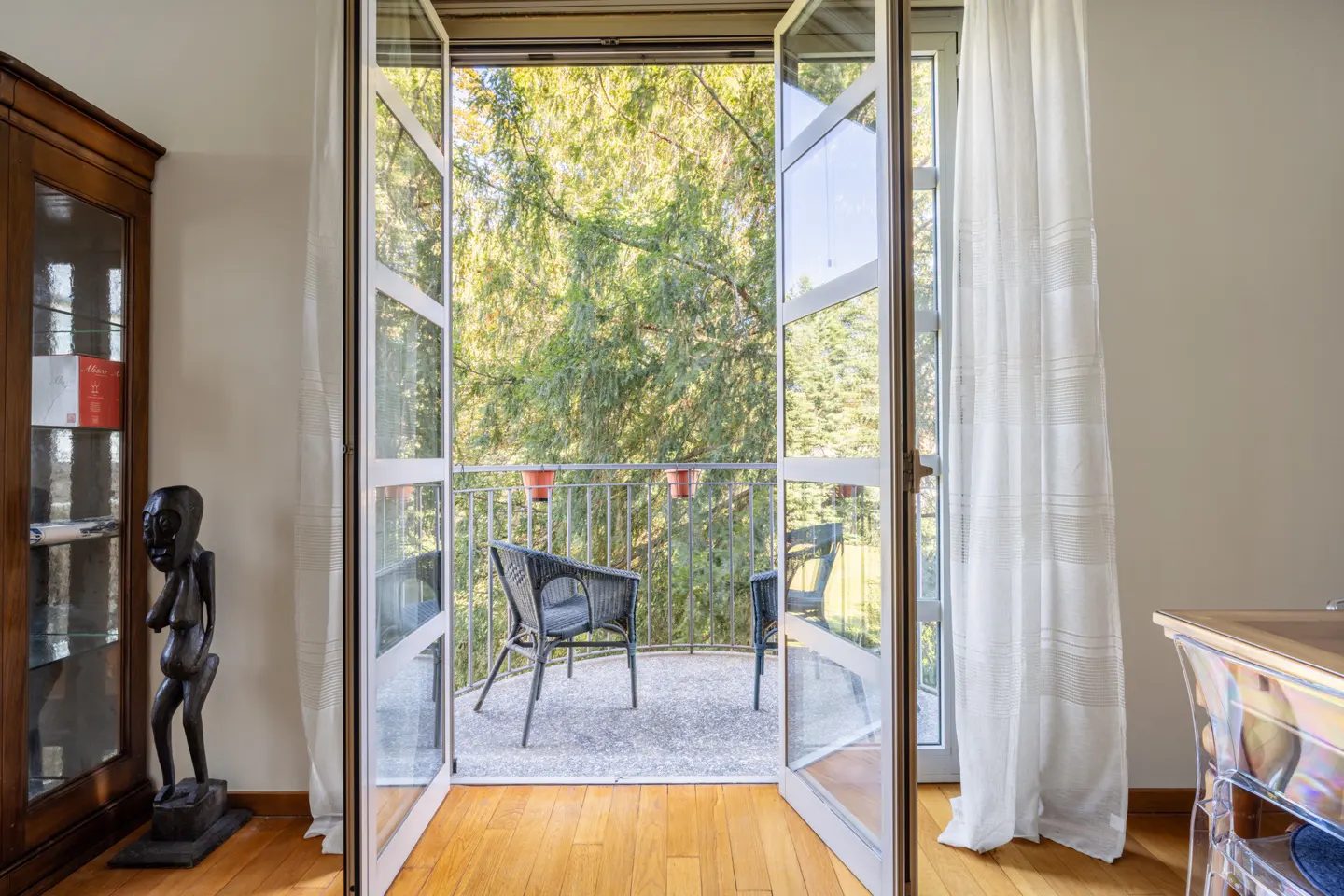 Open French doors lead to a balcony with chairs and greenery. A wooden floor, cabinet, and statue are visible inside.
