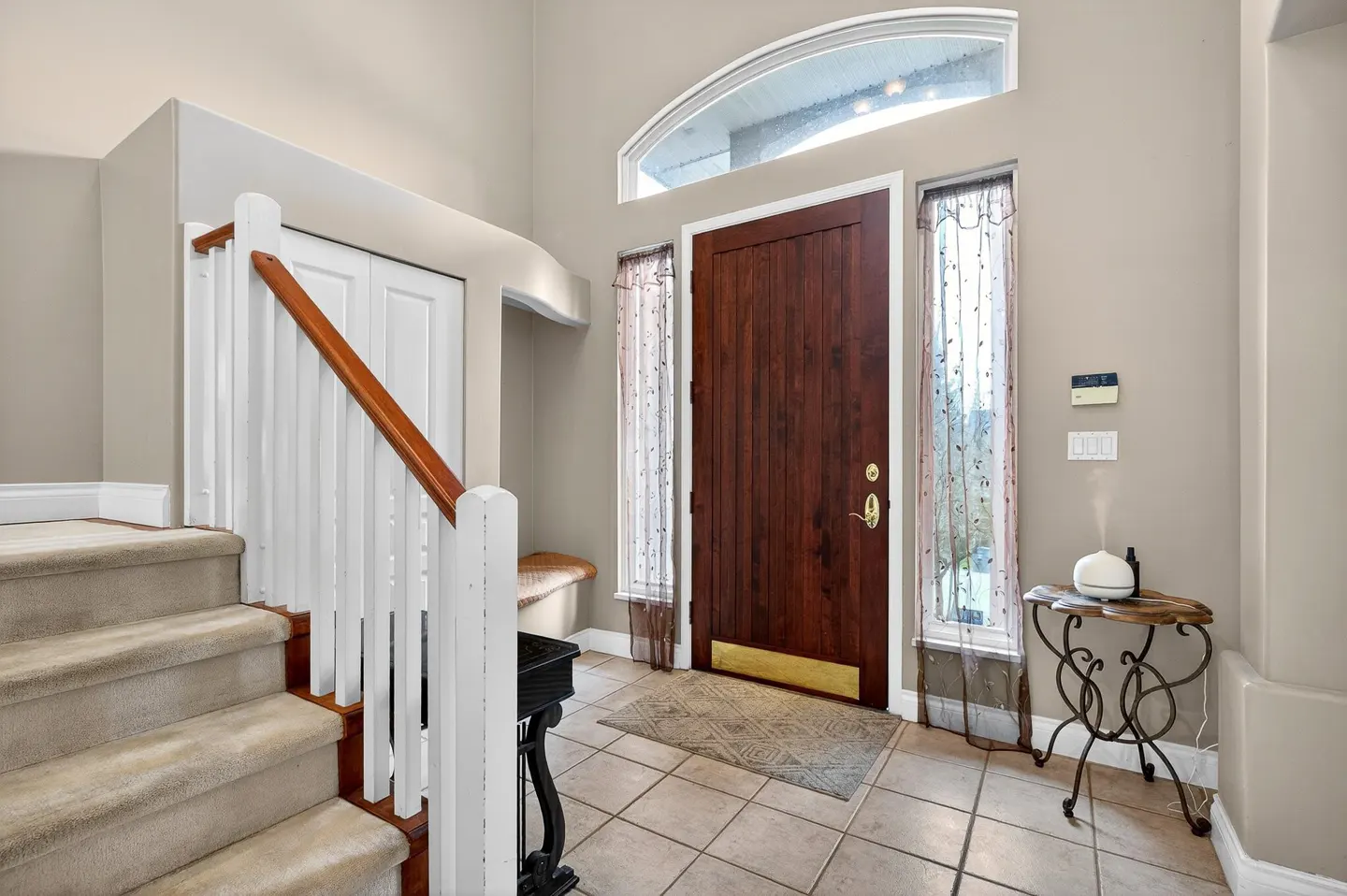 Entryway with a dark wood door, arched transom window, tiled floor, carpeted stairs, and a small side table with a diffuser.