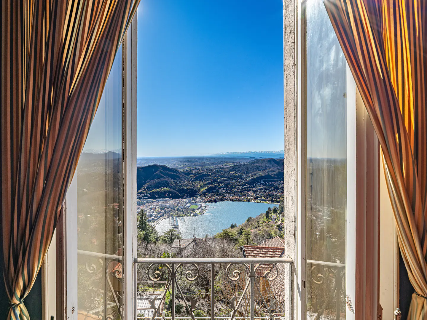 View from an open window with striped curtains, overlooking a lake, mountains, and a clear blue sky. A decorative wrought iron balcony is visible.