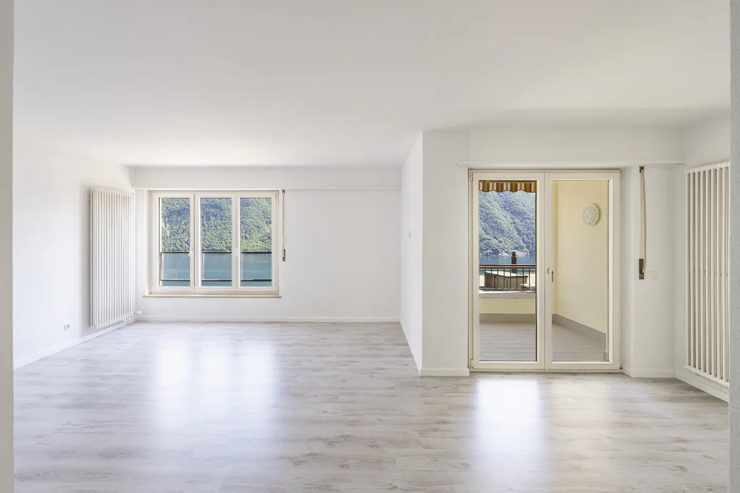 Bright, empty room with white walls, light wood floors, vertical radiators, and a balcony overlooking a lake and mountains.