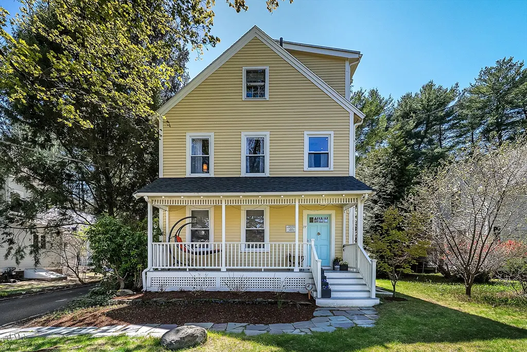 Two-story yellow house with a porch, white railings, and a light blue door. Green trees surround the house.