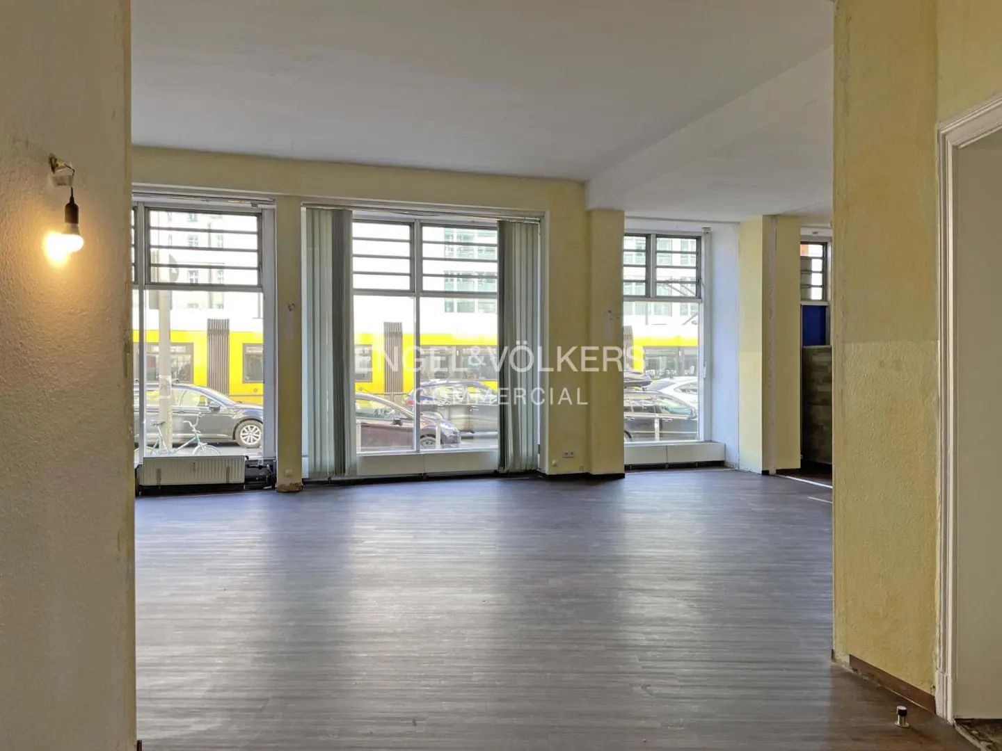 Empty commercial space with wood floors, large windows, and yellow walls. A street view with cars and a yellow train is visible through the windows.
