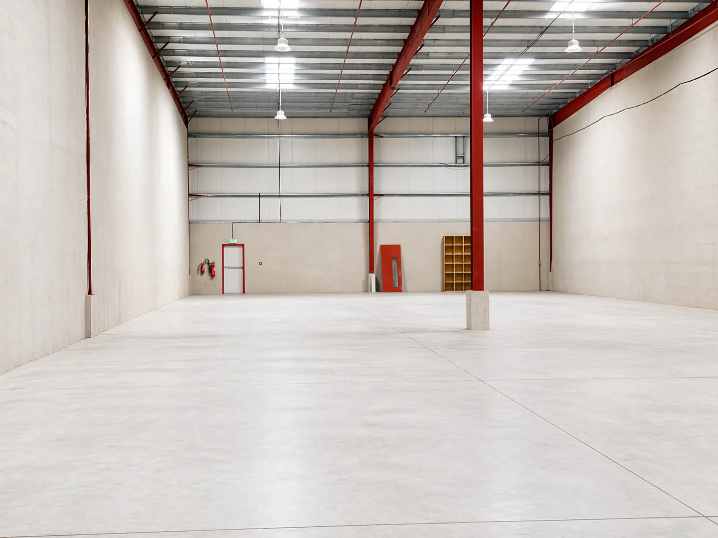 Empty warehouse interior with concrete floor, white walls, and red steel support beams. An emergency exit door and fire extinguisher are visible.