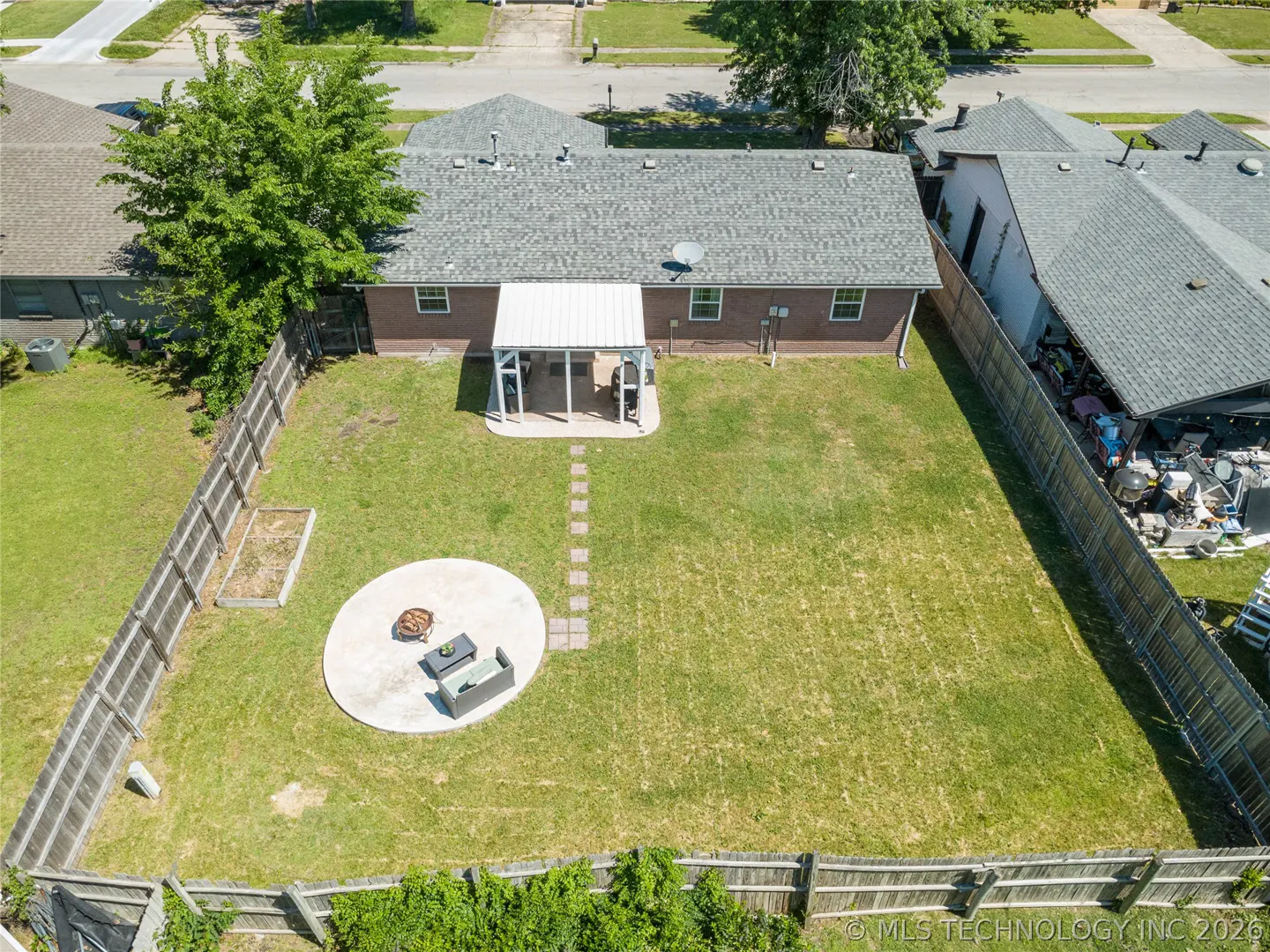 Aerial view of a fenced backyard with green grass, a patio with furniture, and a covered porch attached to a brick house.