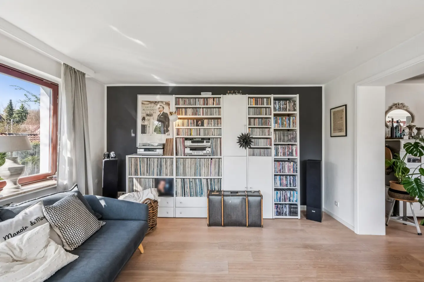 Living room with a blue sofa, wood floors, and a large white shelving unit filled with records, CDs, and DVDs. A vintage suitcase sits in front of the shelves.