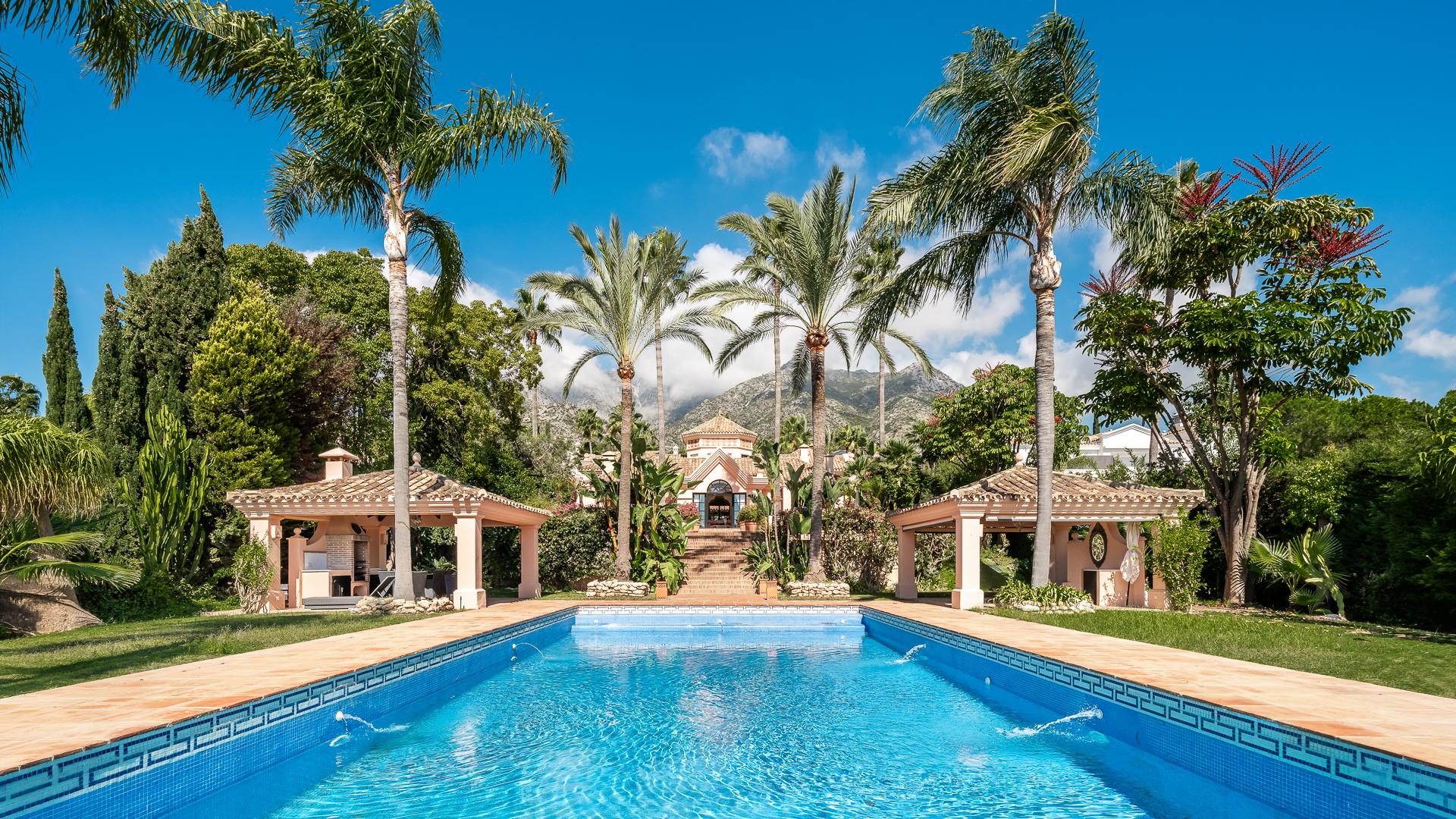 A rectangular blue swimming pool with fountains, flanked by two gazebos, palm trees, and a villa against a mountain backdrop. [AI-generated text]