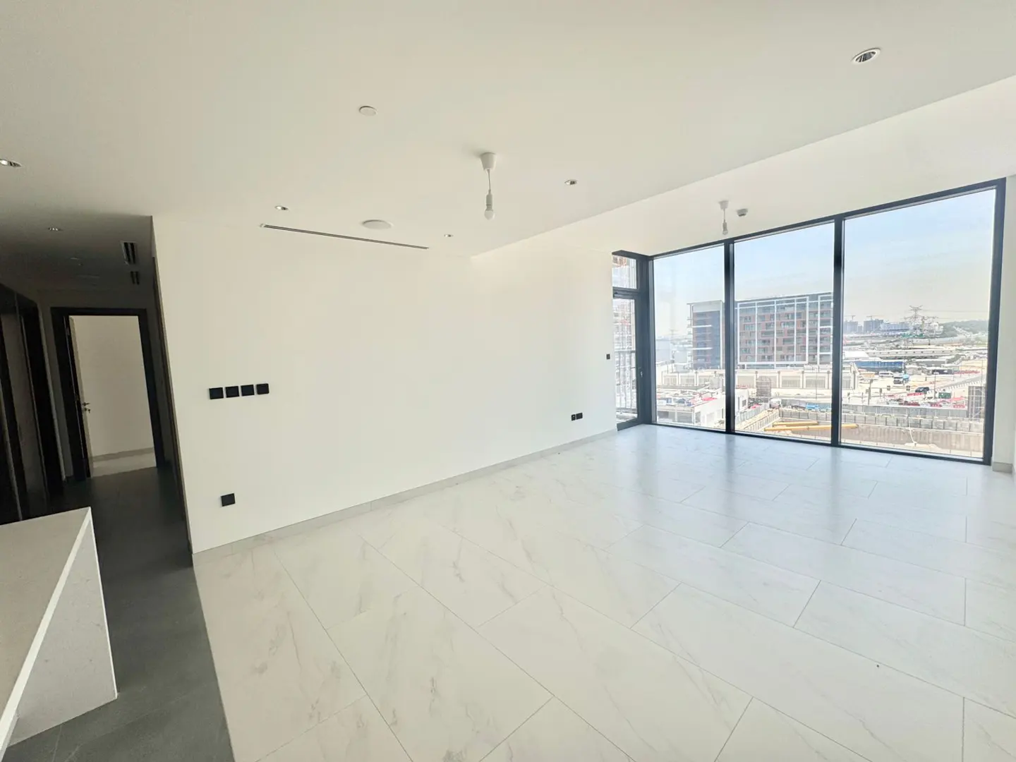 Bright, empty room with white walls and marble floors. Large windows offer a city view. A dark doorway is visible on the left.