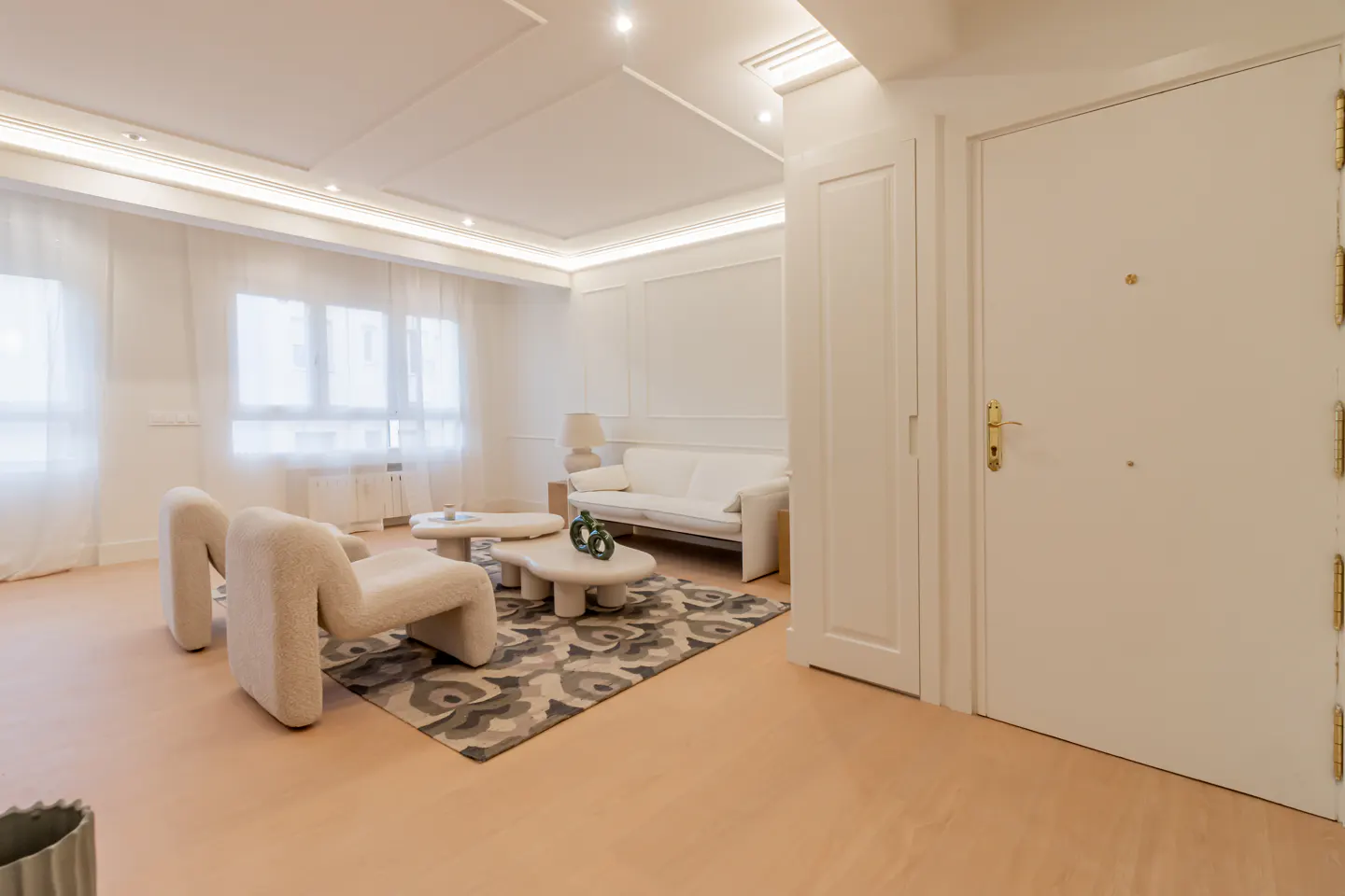 Bright living room with white walls, modern cream armchairs, a sofa, and a patterned rug on light wood flooring.