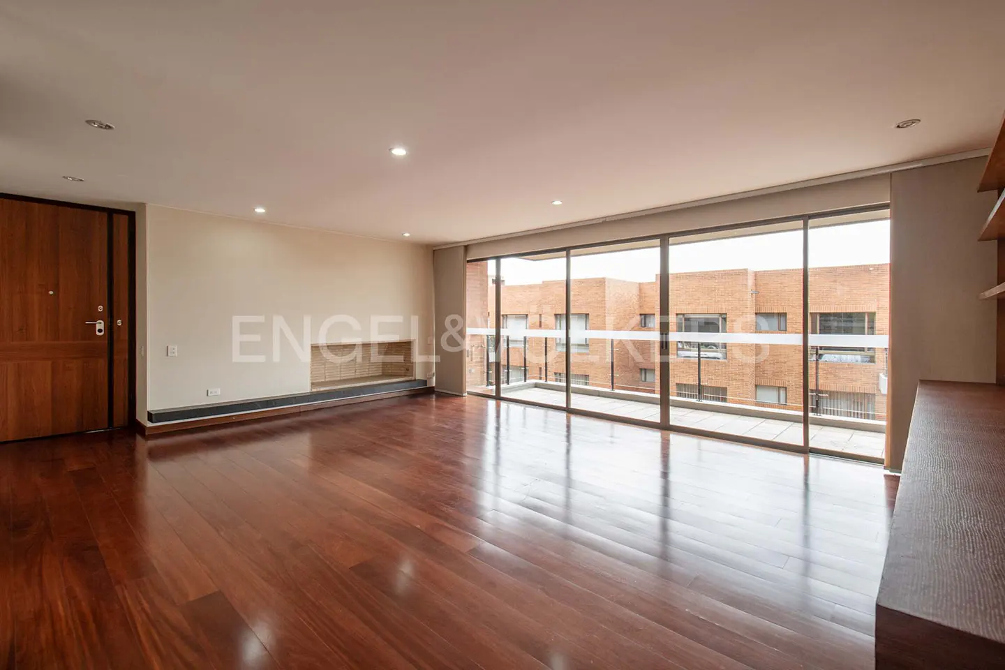 Empty living room with dark wood floors, a large window showing a brick building, and a wooden door on the left.