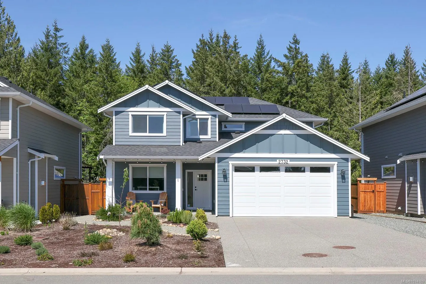 Two-story blue house with white trim, a white garage door, and solar panels on the roof. Trees in the background.